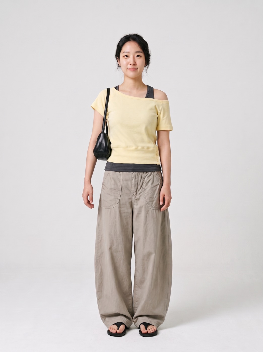 Saran padu padan Style Sketch: atasan satu bahu warna kuning mentega dilapis di atas tank top abu-abu gelap, dengan celana kaki lebar warna cokelat muda, sandal hitam, dan tas bahu hitam kecil.